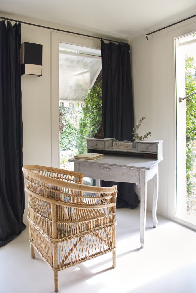 Chambre aux teintes claires avec rideaux foncés, fauteuil en rotin et mobilier patiné sur un sol en béton ciré off-White.