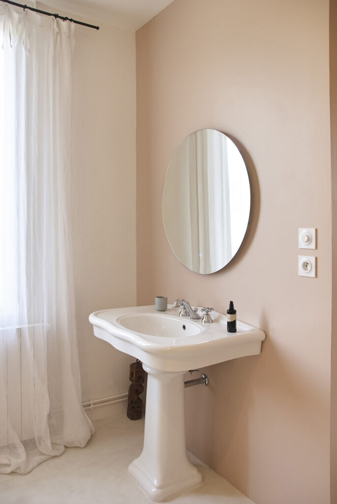 Salle de bain aux teintes rose poudrée de Farrow & Ball avec lavabo ancien sur colonne et grand miroir rond.