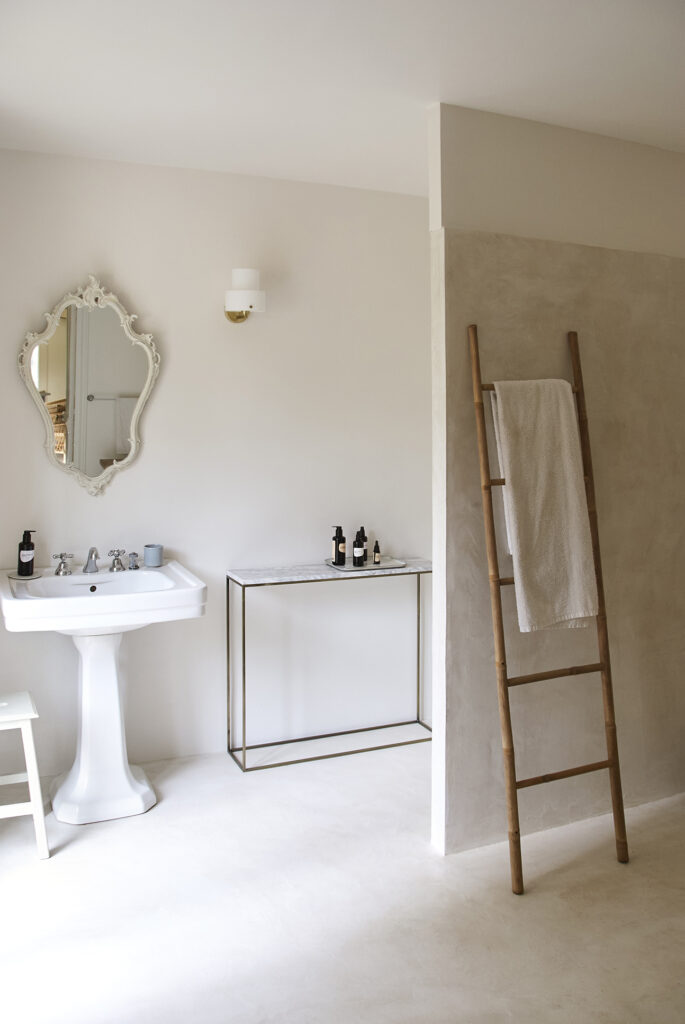 Salle de bain avec lavabo et colonne d'origine, miroir ancien et béton ciré aux teintes douces habillant le sol et les murs.