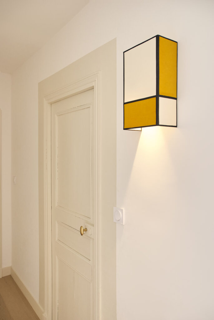 Porte ancienne, encadrement graphique et applique murale bicolore jaune safran et ivoire sur mur clair.